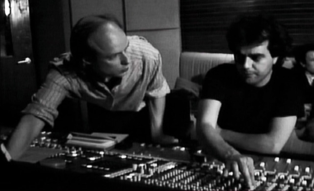 Brian Eno and Daniel Lanois sat at a studio mixing desk. Brian is stood up and adjusting a setting on the desk.
